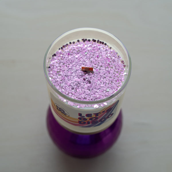 A top-down view of Everlong Candle Co.'s Let's Go Disco candle, showcasing its vibrant purple eco-glitter covering the coconut-soy wax surface. Glitter is sustainably made from eucalyptus trees. Features: crackling wood wick, clear glass jar reveals its 70s-inspired label with bold typography and wavy stripes. Beneath the candle, a purple disco ball adds playful, nostalgic touch.