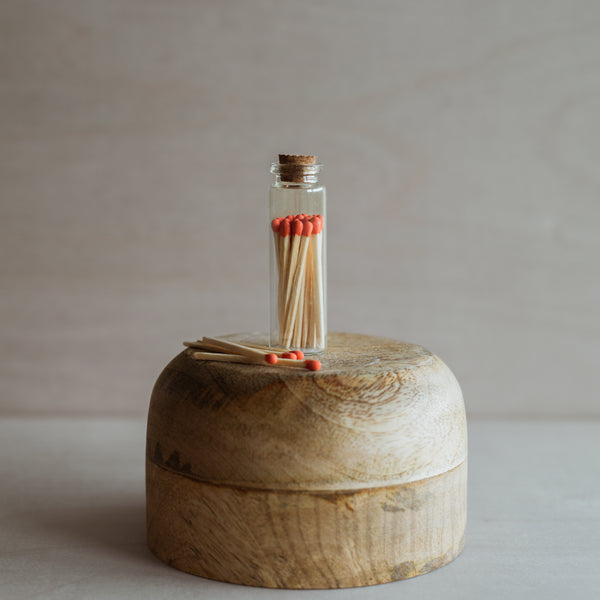 Small, clear glass bottle with a cork lid, filled with orange tipped matchsticks. The bottle is displayed atop a wood stand, four matches lay next to the bottle