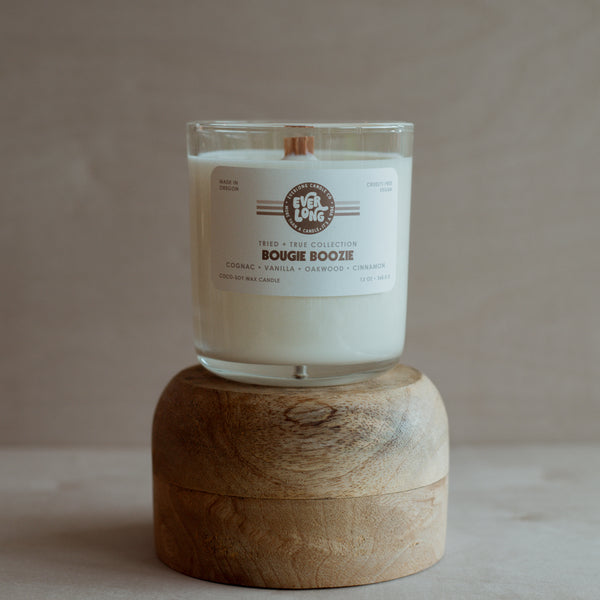 Everlong "Bougie Boozie" candle (13oz) on a wooden stand. Clear glass jar, white coco-soy wax, wood wick. White label with brown stripes. "Bougie Boozie" in large font, scent notes: cognac, vanilla, oakwood, cinnamon.