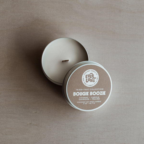 An open "Bougie Boozie" candle tin (2 oz). Cognac, vanilla, oakwood, praline scent. White coco-soy wax, wood wick. Round, brown label with white retro logo and text.
