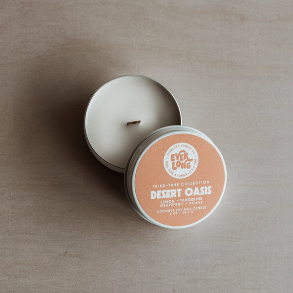 White 2 oz. tin candle, open lid resting beside it. Round, orange label with white text, Everlong logo and Desert Oasis in bold font. Scent notes: lemon, tangerine, grapefruit, agave. Smooth coconut-soy wax with wood wick.