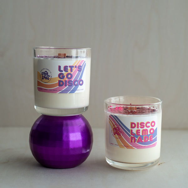 Everlong Candle Co.'s Let's Go Disco and Disco Lemonade candles in clear glass jars are displayed together. Let's Go Disco sits atop a purple disco ball, Disco Lemonade, rests beside it. Both candles feature a crackling wooden wick, and glitter