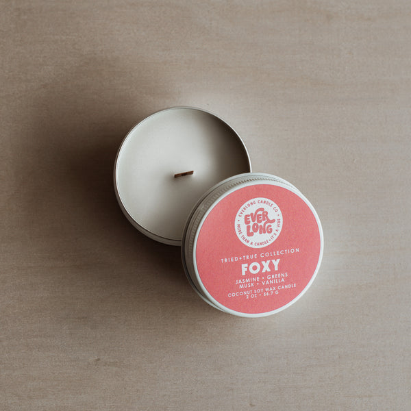 Everlong "Foxy" (2 oz.) candle tin, lid off, on a wooden surface. White coco-soy wax, wood wick. Round, coral colored label "Foxy" in large font, scent notes: jasmine, greens, vanilla, musk.