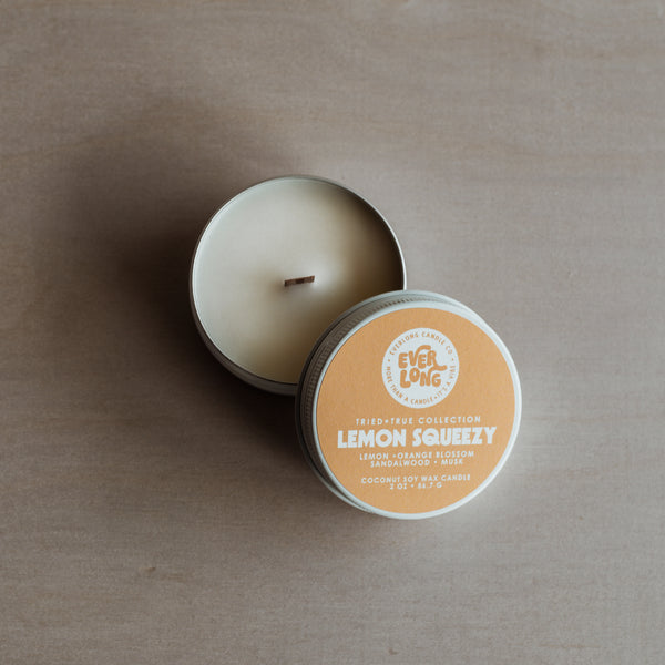 Open Everlong "Lemon Squeezy" candle tin (2 oz) on a wooden surface. White coco-soy wax, wooden wick. Round, yellow label with black text, "Lemon Squeezy" in large font, scent notes: lemon, orange blossom, sandalwood, musk.