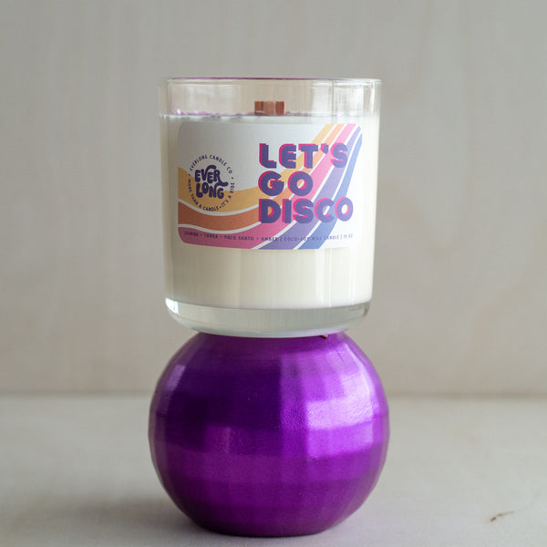 Everlong Candle Co.'s Let's Go Disco 13 oz hand-poured scented candle in a clear glass jar with a crackling wooden wick. Features a contemporary 70s-inspired label with bold retro typography and wavy stripes. Topped with sustainable, biodegradable purple glitter for a touch of disco magic. This vegan, cruelty-free coconut-soy wax candle fills the air with rich, warm notes of jasmine, tonka, palo santo, and amber. Burns for 65+ hours. Free from paraffin, phthalates, parabens, and carcinogens.