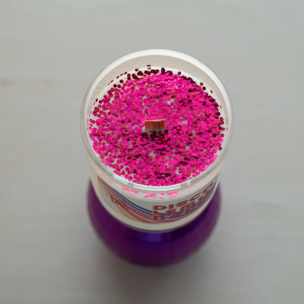 A top-down view of Everlong Candle Co.'s Disco Lemonade candle, showcasing its vibrant pink biodegradable glitter covering the smooth coconut-soy wax surface. Non-toxic. Made from eucalyptus trees. Features: Clear glass jar, wood wick, 70s-inspired label with bold typography and wavy stripes. Beneath the candle, a purple disco ball.
