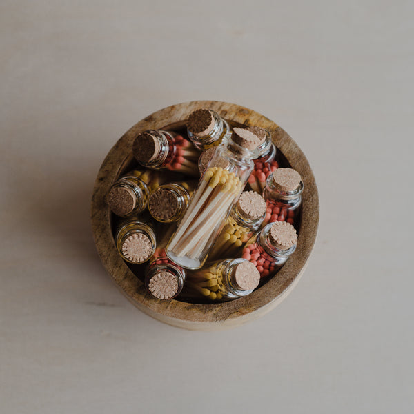 Wood bowl filled with little clear glass jars filled with orange and yellow tipped matches, topped with a cork lid