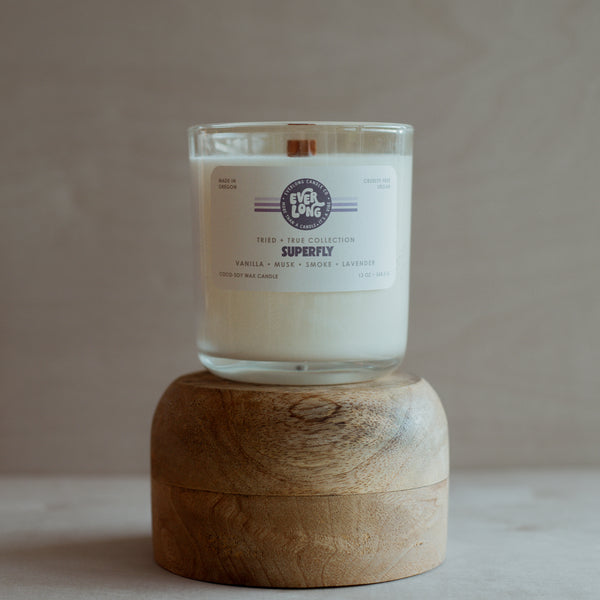 Clear glass jar candle on a wooden stand. The label reads "Everlong Superfly" with vanilla, musk, smoke, and lavender scent notes. A wooden wick is visible in the white candle wax. "Made in Oregon" is also on the label.