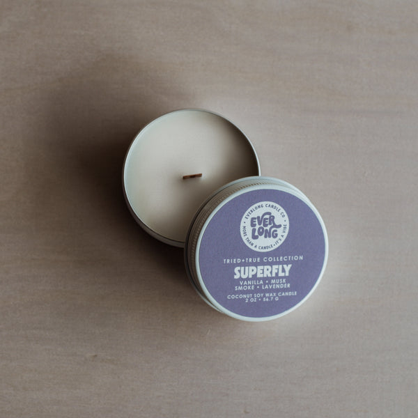 Round, white tin candle (2 oz), lid resting beside it. Purple label with Everlong Logo and Superfly in bold font. Scent notes: vanilla, musk, smoke, lavender. Wood wick visible in white wax.