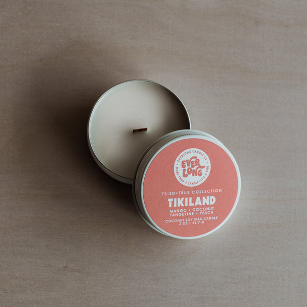 White tin candle, open lid resting beside it. Warm pink label with Everlong and Tikiland in bold font. Scent notes: mango, coconut, tangerine, peach. Wooden wick visible in white wax.