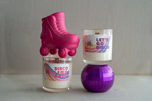 Two candles in clear glass jars filled with white coconut-soy wax, wood wicks, and pink and purple eco-glitter sprinkled on top. Accompanies by a ceramic roller skate and a purple disco ball.