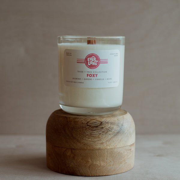 Clear glass jar candle on a wooden stand. White label with warm pink stripes, "Everlong Foxy" in bold font. Scent notes: jasmine, greens, vanilla, musk.  Wooden wick visible in white wax. "Made in Oregon.