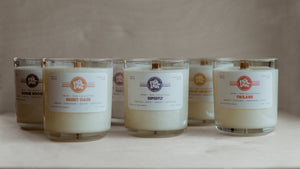 Six 13 oz clear glass candles without lids, arranged in two rows on a wooden surface. All have white labels printed with bright colors, showcasing the Everlong logo and scent names: "Bougie Boozie," "Foxy," "Lemon Squeezy," "Desert Oasis," "Superfly," and "Tikiland." All have white wax and wood wicks.